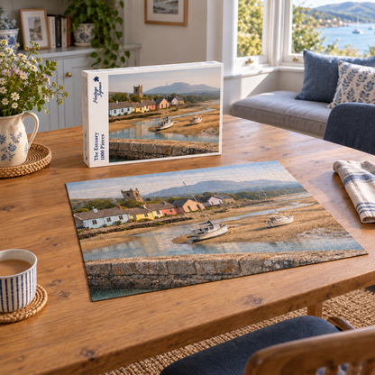 The Estuary 1000 piece jigsaw box and completed puzzle on an oak table, showing yachts grounded on the sand at low tide below pastel cottages, a church tower and distant mountains