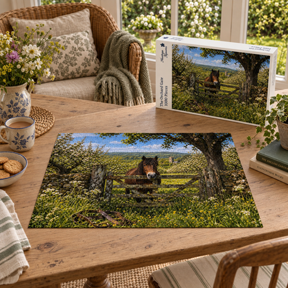 The Orchard Gate 1000 piece jigsaw box and completed puzzle on an oak table, showing a bay pony leaning over a five bar gate in a blossoming English orchard